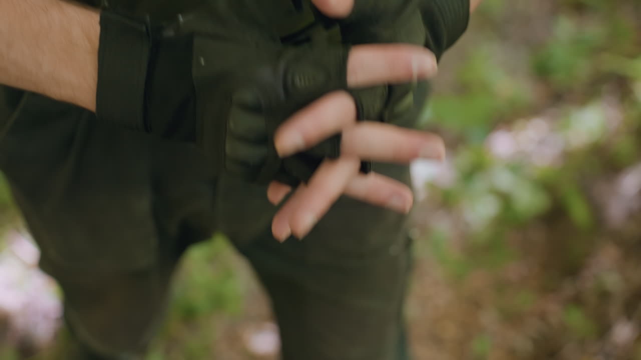 soldier wearing fingerless glove on left hand and putting on rubber wool glove on right, standing ready for fight in sunlit forest clearing, belt and cargo pants visible under dappled canopy