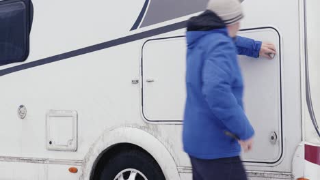 una persona con una chaqueta azul abre la puerta de carga lateral de una caravana de barro blanco durante una aventura de invierno