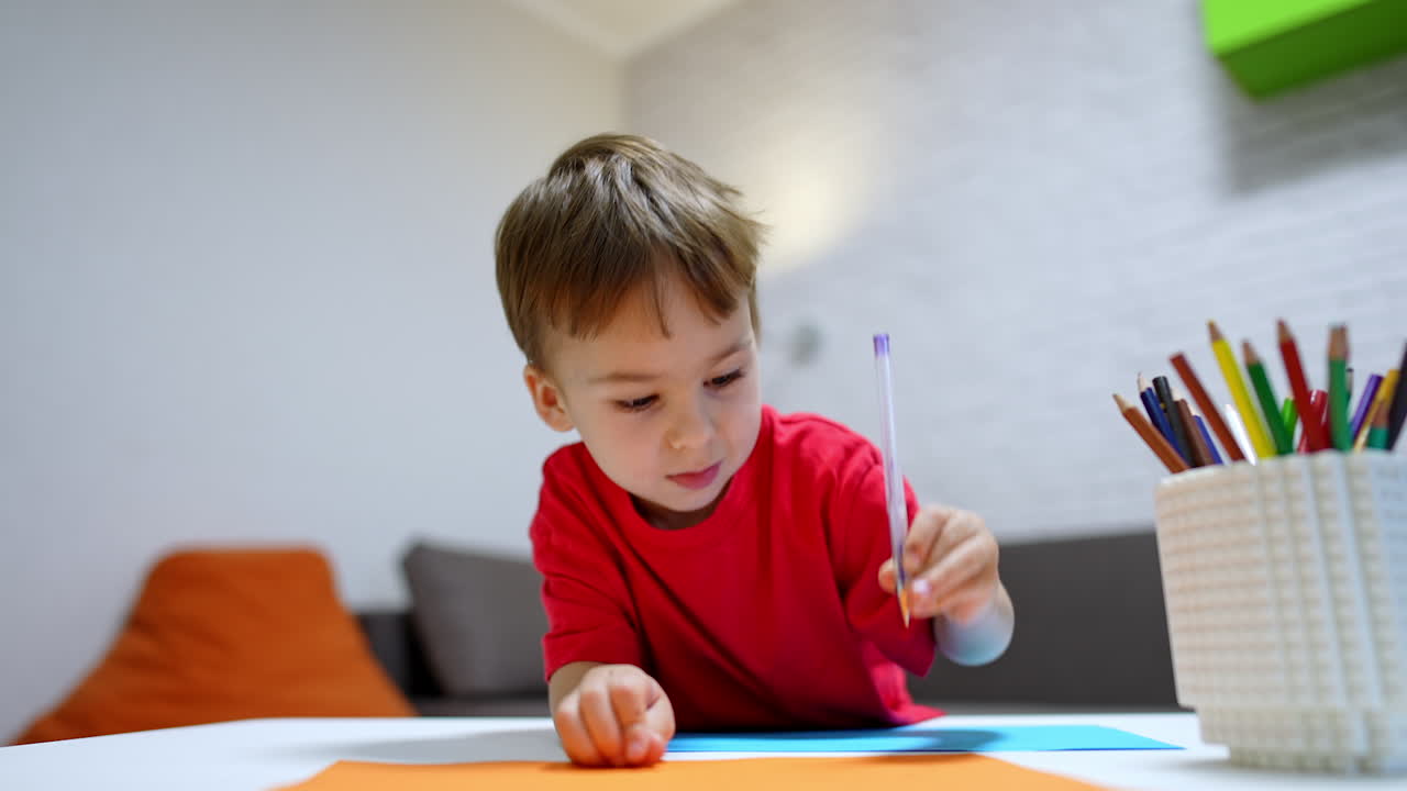 Caucasian toddler kid in red t-shirt draws on the desk. Focused baby boy drawing and chooses a pencil from a cup. Low angle view.