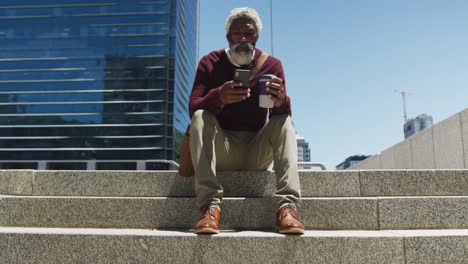 African-american-senior-man-holding-coffee-cup-using-smartphone-while-sitting-on-the-stairs-at-corpo