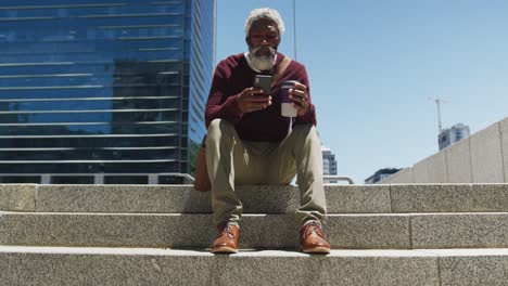 hombre mayor afroamericano sosteniendo una taza de café usando un teléfono inteligente mientras estaba sentado en las escaleras en el cuerpo