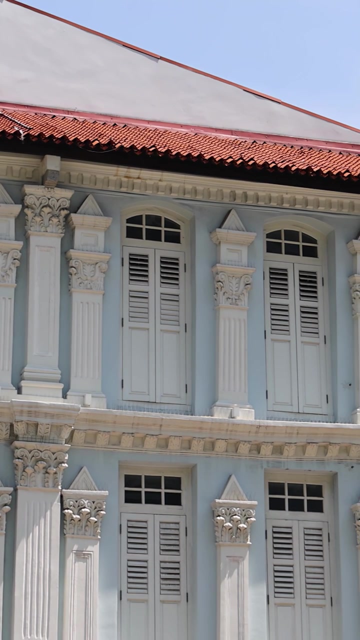 La telecamera si muove su case coloniali a schiera blu pastello con colonne ornate e finestre con persiane sotto la luce del sole, terminando su moderni edifici cittadini e insegne.