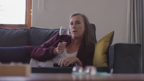 woman watching tv and drinking wine while laying on the couch at home