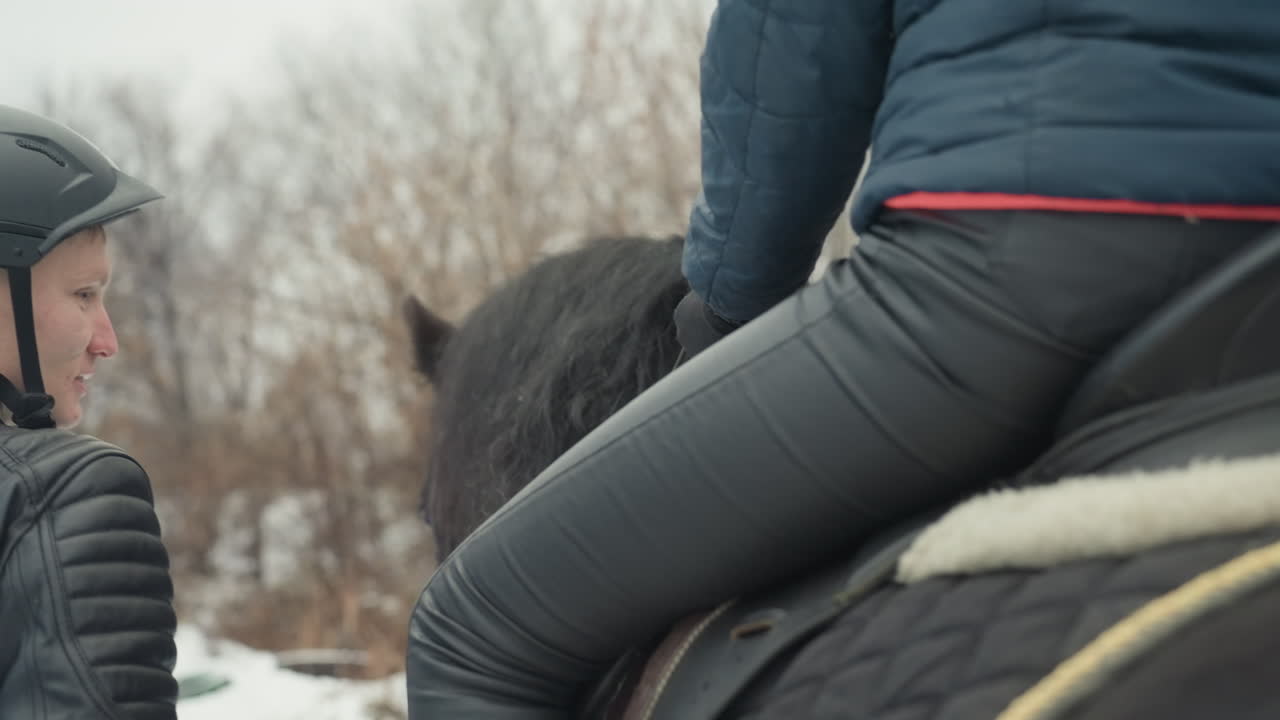 雪原で馬に乗る騎手を補助するタンデム乗馬インストラクター。ヘルメットをかぶったコーチが鐙に足を導き、鞍に手を置いています。バランスと姿勢に関する密接な指導と、革製馬具のディテールが見えます。