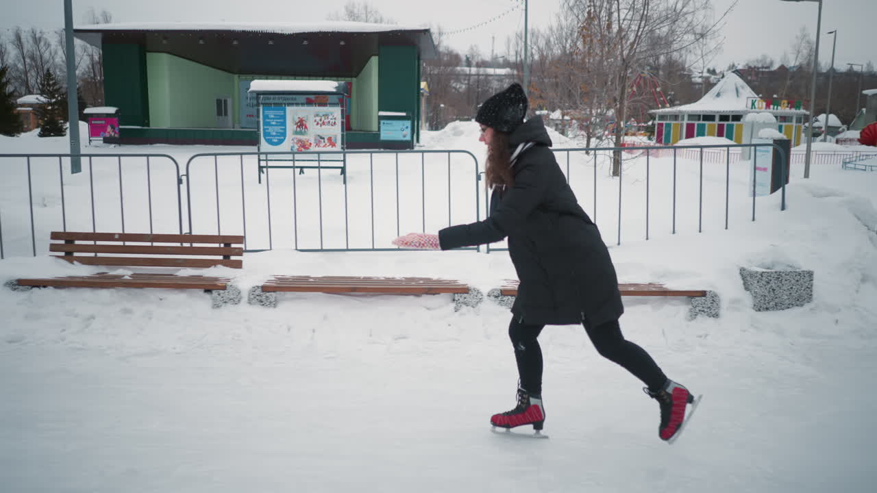 Lady skating outdoors on frozen ice rink during winter, dressed in black coat, red skates, warm hat, colorful gloves, and scarf, practicing movement near snowy benches and park structures surrounded by snow