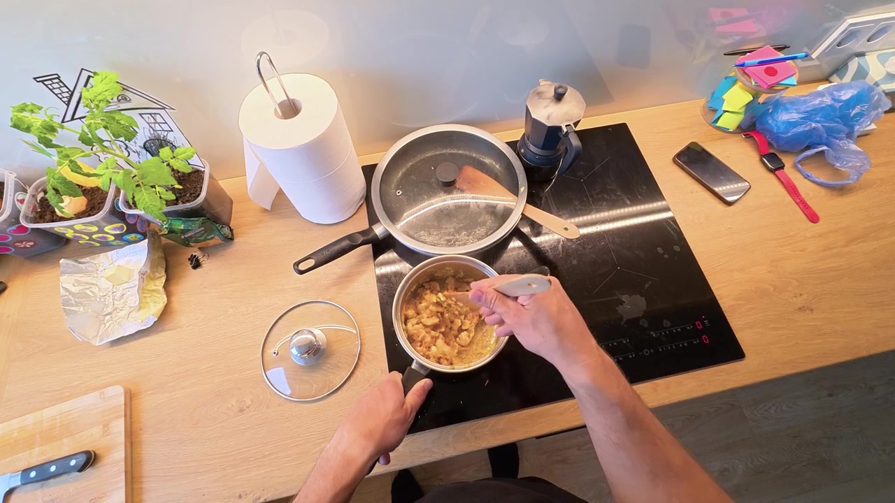 Ripresa POV dall'alto di una persona che mescola cibo in una casseruola su un piano cottura a induzione, circondata da utensili da cucina, erbe fresche e oggetti vivaci sul piano di lavoro sotto luce naturale.