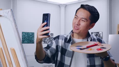 primer plano de un artista masculino asiático haciendo una llamada de video en un teléfono inteligente mientras pinta en lienzo con pinturas al óleo y pincel en la galería