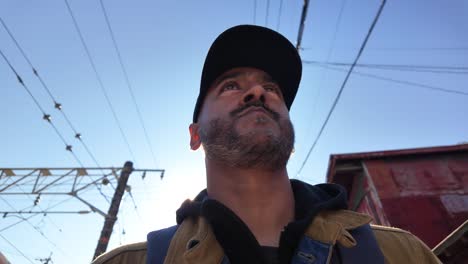man in baseball cap gazing upward, with railway wires and blue sky background.