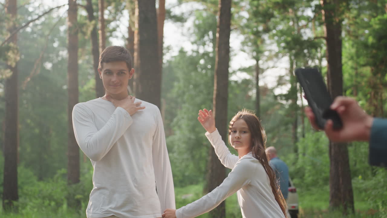 Young girl posing beside big brother while mother takes photo with phone in sunny forest, family enjoying cheerful outdoor moment surrounded by trees and natural light creating emotional connection