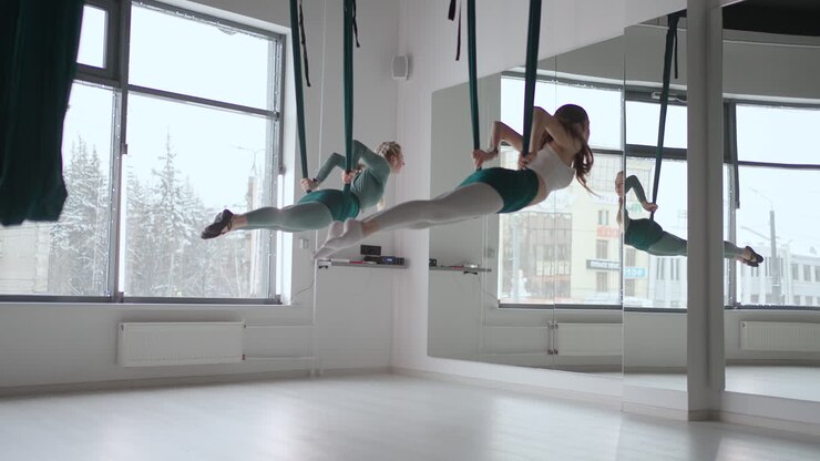 Due giovani donne yogi praticano yoga aereo in amache verdi in una palestra. Belle donne si allenano in classe eseguendo aero yoga. Variazione della posa della Piramide Parsvottanasana.