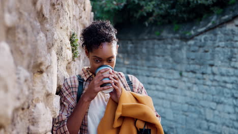 Millennial-woman-with-short-curly-hair-wearing-plaid-shirt-leaning-on-stone-wall-in-the-street-drinking-a-takeaway-coffee,-close-up