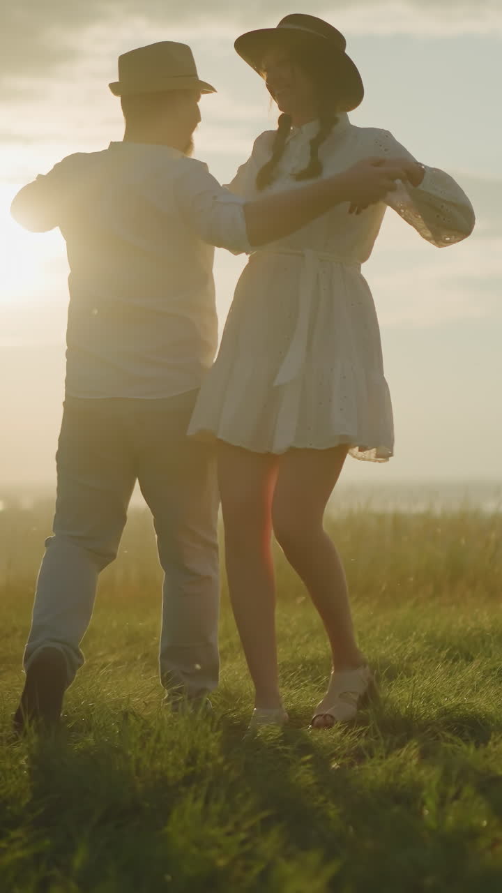 빛이 빛나는 잔디 언덕에서 부드럽게 춤을 추는 두 마리의 러브버드의 만적 인 장면, 함께 사랑의 순간을 공유합니다. 색 셔츠, 모자, 청바지를 입은 남자는 색 드레스를 입은 아내를 들고 있습니다.