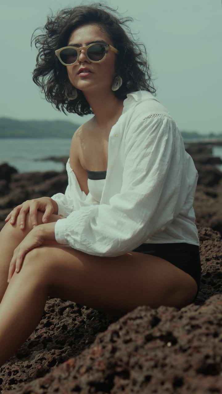 Vertical shot of a South Asian woman in sunglasses and white shirt sitting on rocks by the sea