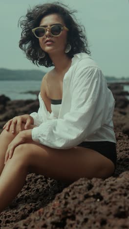 vertical shot of a south asian woman in sunglasses and white shirt sitting on rocks by the sea