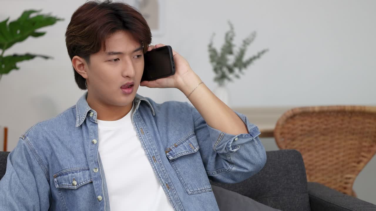 ชายหนุ่มชาวเอเชียสวมเสื้อเชิ้ตยีนส์กำลังคุยโทรศัพท์มือถืออย่างจริงจังภายในอาคาร