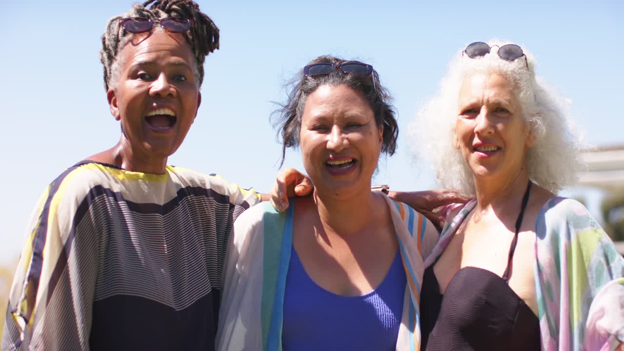 Amigas diversas dándose cuenta de la cámara, riendo, entrelazando los brazos, saludando con gafas de sol en el paseo marítimo.