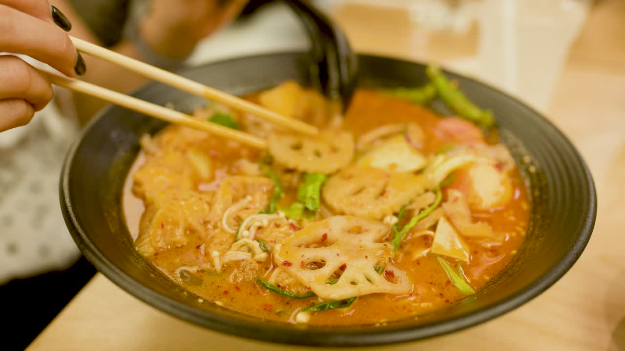 Hand uses chopsticks to lift noodles from spicy Asian soup in warm, indoor restaurant lighting
