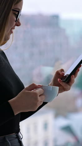 pretty girl with a phone in business office. long haired young woman in glasses drinks coffee near the large window with city background and looks into the phone. vertical video