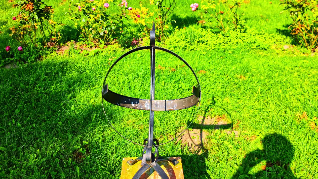 Close-up view of an old metal sundial showing Roman numerals and a shadow on green grass in a sunny Jaunpils garden - push in to close-up