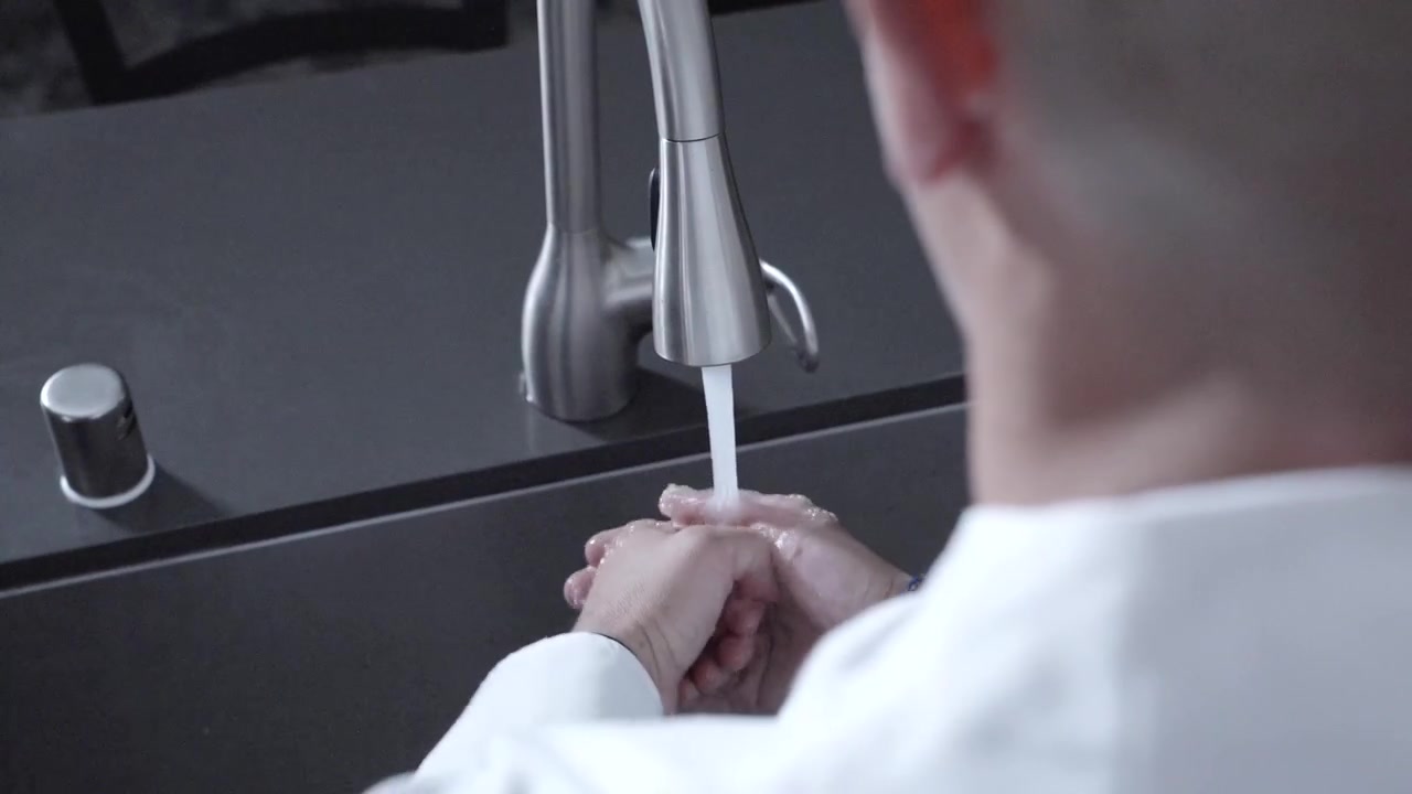 Inquadratura statica da sopra la spalla di uno chef che si lava le mani in una cucina pulita, enfatizzando le pratiche di igiene e sicurezza alimentare essenziali per la preparazione culinaria.