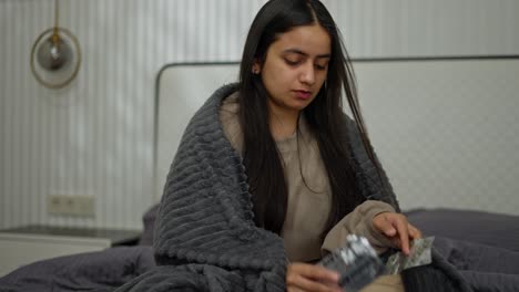 una chica morena con un aspecto enfermo elige qué pastillas debe tomar saca una y la lava con agua en un apartamento moderno durante una enfermedad en el dormitorio