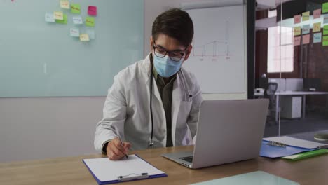 Asian-male-doctor-wearing-mask-sitting-at-desk-in-meeting-room-making-notes