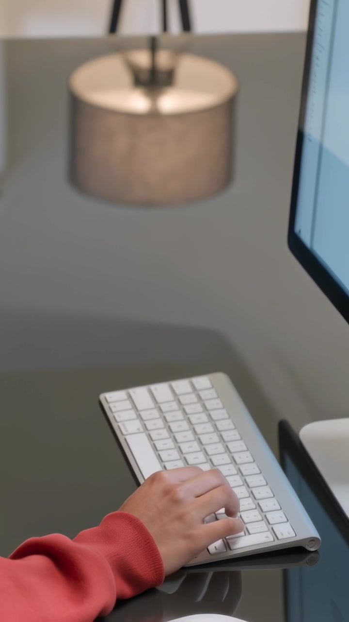 縦型動画：赤いトップスを着た女性がモニターのテキストを読み、キーボードでタイピングしている。コピースペースあり。