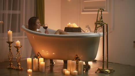 a young beautiful caucasian brunette lies in the bathroom by candlelight in a pleasant evening atmosphere resting from stress and relaxing drinking red wine from a glass