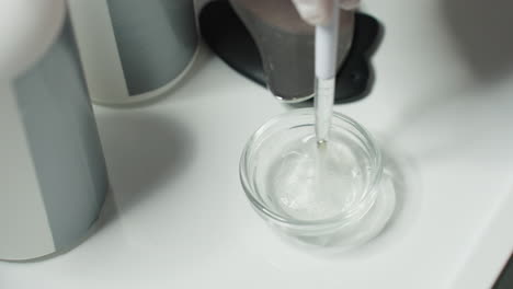 hand on protective glove stirring clear facial gel in glass container on white spa tray under soft light highlighting viscous texture and professional skincare ritual details precision