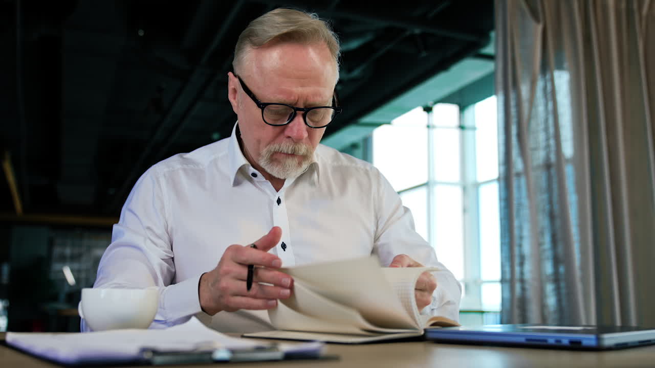 Male wearing glasses sits at desk turning pages of his paper notebook. Man reads something out loud grimacing. Low angle view.