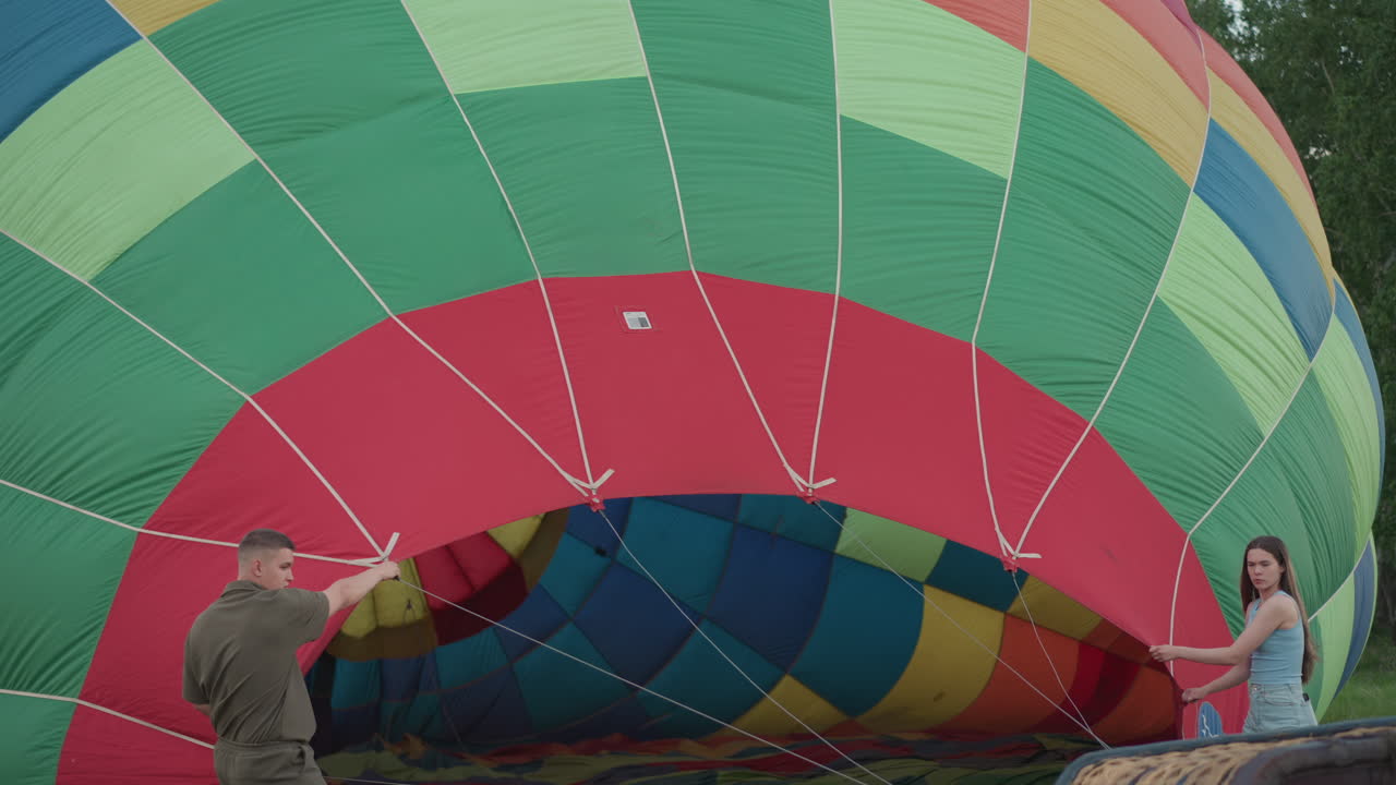 couple holding hot air balloon envelope in open grassy field at golden hour preparing for inflation with gentle breeze rippling colorful nylon while crew and equipment stand ready in background