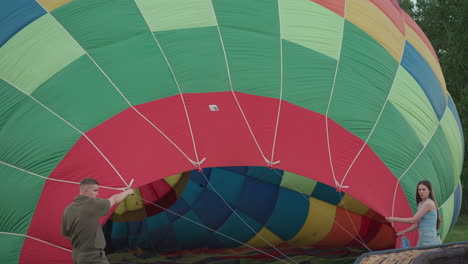 couple holding hot air balloon envelope in open grassy field at golden hour preparing for inflation with gentle breeze rippling colorful nylon while crew and equipment stand ready in background