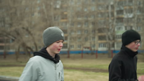 close-up of two boys jogging gradually, with one wearing glasses and a gray beanie, as they jog, a man in a brown beanie speeds past them on the track, with a blurred background