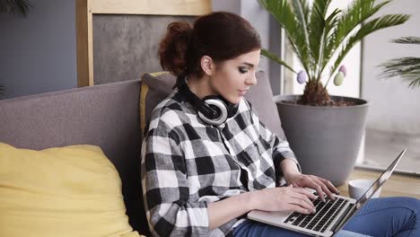 A-trendy-brunette-girl-with-a-laptop-in-her-hands-flops-on-the-sofa-and-starts-typing.-Hedphones-on-neck.-Modern-interior