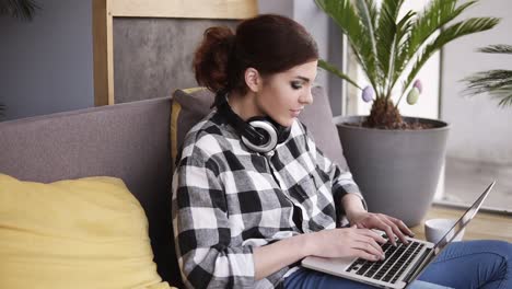 una chica morena de moda con una computadora portátil en las manos se cae en el sofá y comienza a escribir. auriculares en el cuello. interior moderno