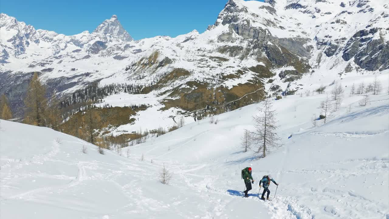 Two hikers trek with snowshoes and backpacks through deep snow in scenic Italian Alps, iconic Matterhorn or Cervino mountain peak under blue sky, Italy. Aerial drone backward, low altitude