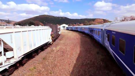 smooth fly in drone shot - establishing blue botswana train , on a sunny hot day with dry grass vegetation and brown soil surrounding the railway line