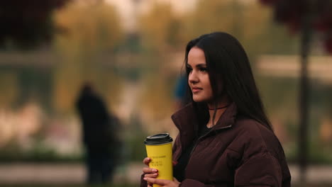 Beautiful-brunette-woman-with-long-hair-walks-in-a-park-holding-a-cup-of-coffee