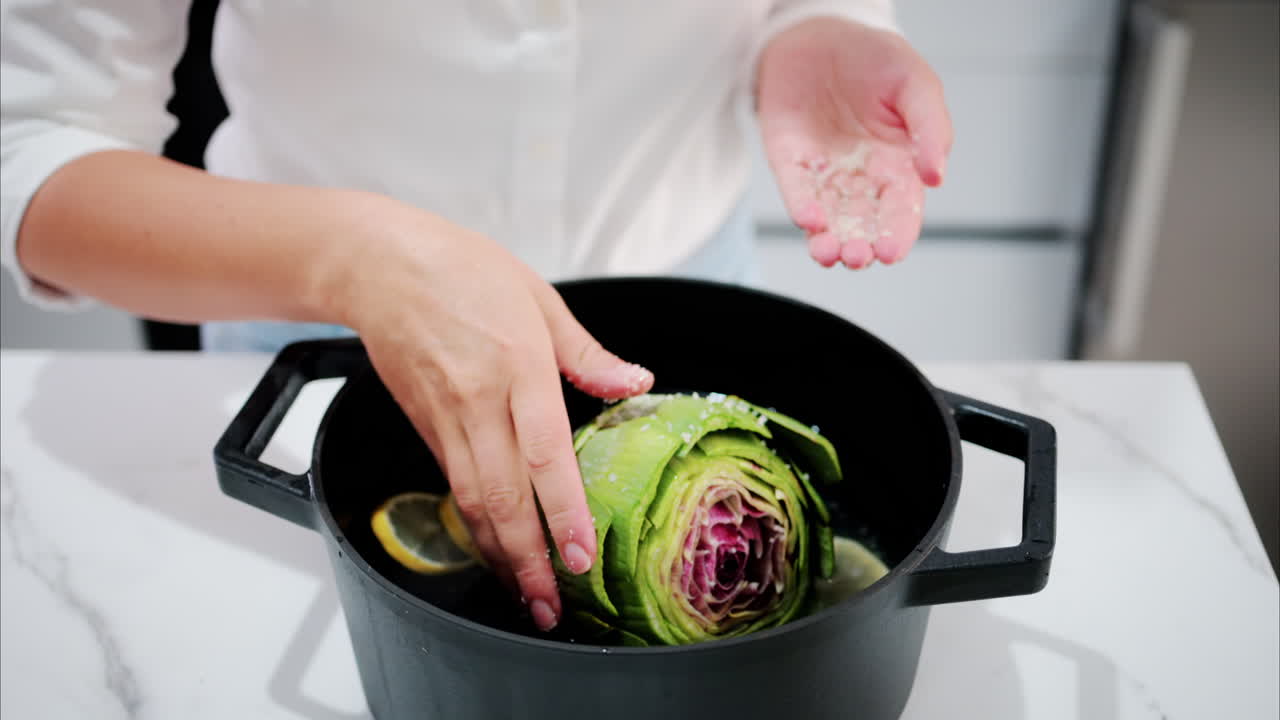 Woman pouring salt over an artichoke in a pot with water and lemons