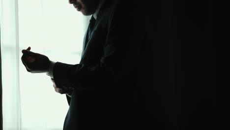 confident businessman in classic blue suit buttoning or adjust cufflinks near window in hotel room at the morning. handsome man wearing a nice suit on wedding day.