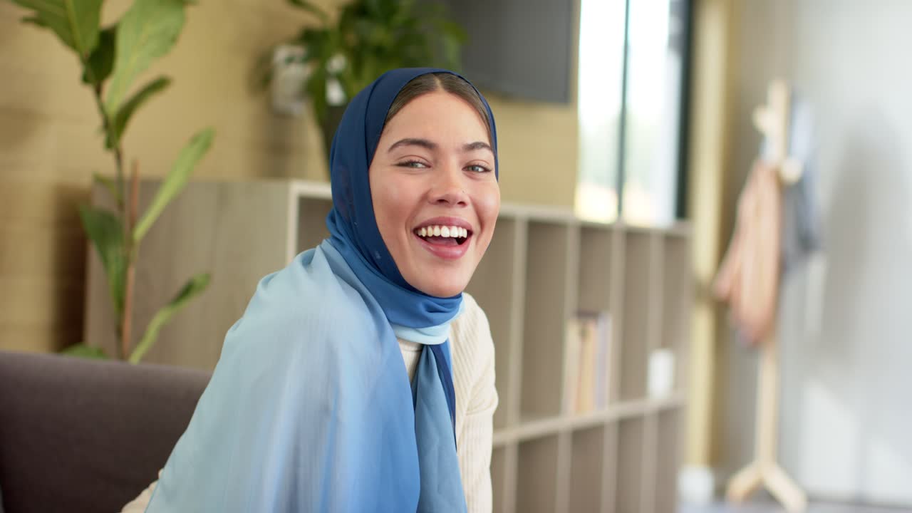 Wanita India memperhatikan kamera saat duduk di rumah di sofa abu-abu, tersenyum lalu tertawa alami.