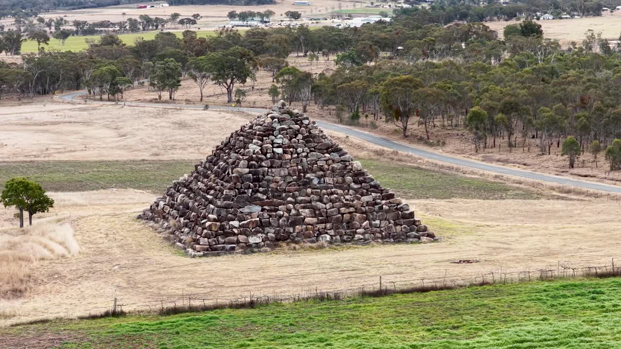 Drone camera circles a large stone pyramid structure in a dry, grassy field with scattered trees under soft daylight in Ballandean, Queensland