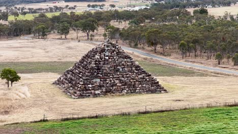 drone camera circles a large stone pyramid structure in a dry, grassy field with scattered trees under soft daylight in ballandean, queensland