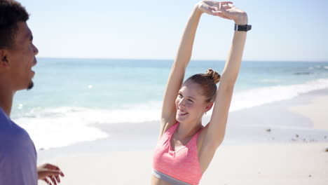 Young-Caucasian-woman-stretches-on-a-sunny-beach,-guided-by-a-young-Biracial-man