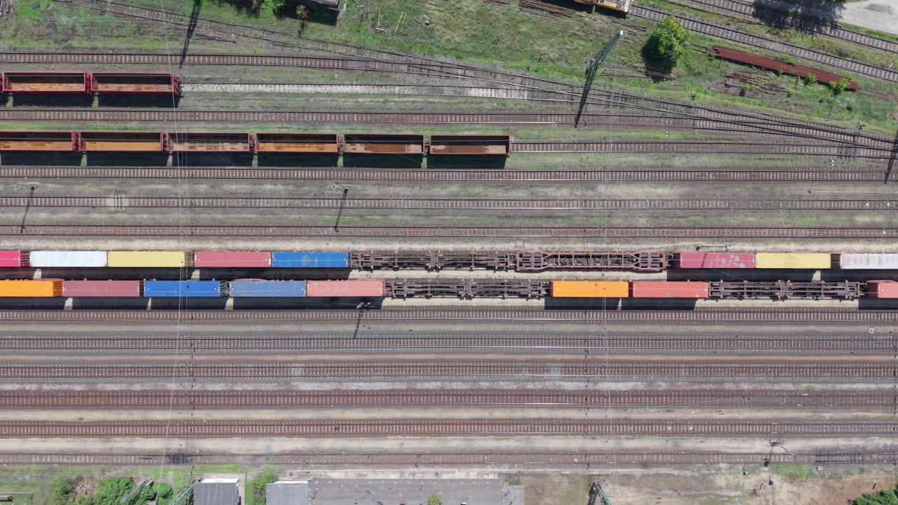 A top-down aerial video showing a freight train loaded with colorful cargo containers in a large railway yard. Multiple train tracks and empty wagons are visible