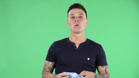 portrait of a handsome young guy looking at camera and removes medical protective mask and breathes air. health protection corona virus concept. green screen