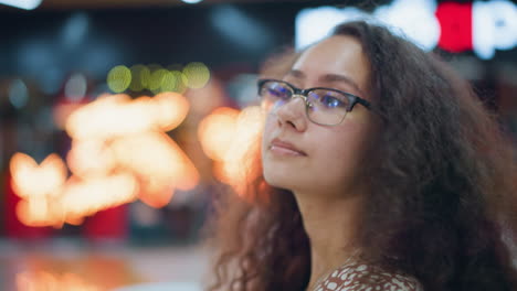 primer plano de una mujer con cabello rizado y gafas sentada en el centro comercial, mirando cálidamente a la distancia con una sonrisa suave, rodeada de luces bokeh suaves y borrosas en el fondo