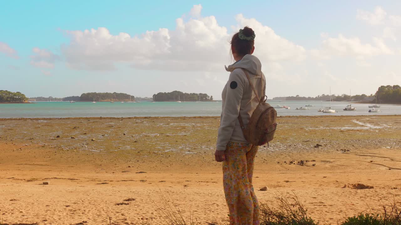 A woman walks along a tranquil shoreline, gazing out at the calm sea and distant boats. The setting is serene, capturing the essence of a sunny day at the beach with gentle waves and soft sand