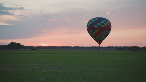hot air balloon with colorful checkerboard canopy descending over green crop field at sunset under pastel sky drifting gently toward horizon in serene twilight landscape