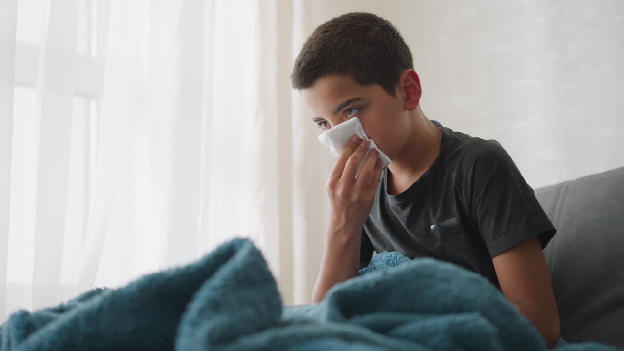 Exhausted young boy sneezing into white napkin covered with blanket, using tissue to wipe his nose while resting on the couch near the window, showing signs of tiredness